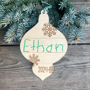 Puede incluir: Un adorno navide&ntilde;o de madera con forma de l&aacute;grima con una cuerda verde para colgar. El adorno est&aacute; decorado con copos de nieve marrones y el nombre "Ethan" escrito con un marcador verde. El a&ntilde;o "2024" tambi&eacute;n est&aacute; escrito en el adorno.