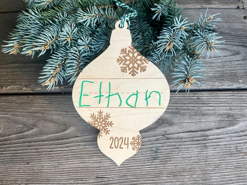 Puede incluir: Un adorno navide&ntilde;o de madera con forma de l&aacute;grima con una cuerda verde para colgar. El adorno est&aacute; decorado con copos de nieve marrones y el nombre "Ethan" escrito con un marcador verde. El a&ntilde;o "2024" tambi&eacute;n est&aacute; escrito en el adorno.