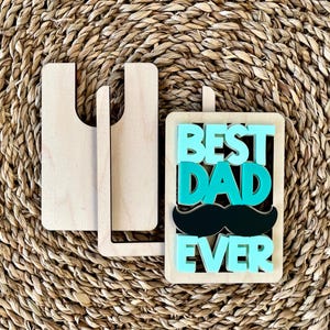 May include: Wooden sign with the text "Best Dad Ever" in teal and black letters. The sign has a black mustache and is propped up on a wooden stand.