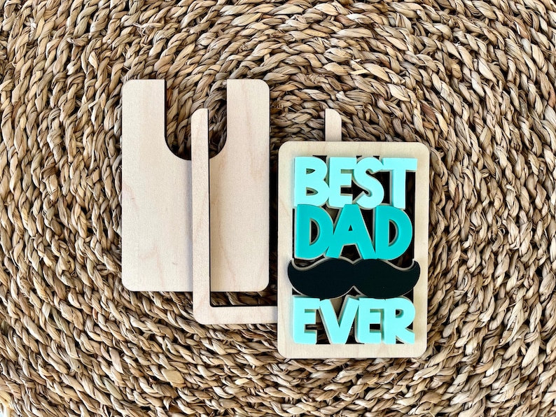 May include: Wooden sign with the text "Best Dad Ever" in teal and black letters. The sign has a black mustache and is propped up on a wooden stand.