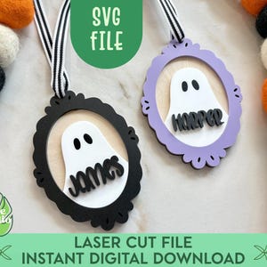 May include: Two Halloween-themed ornaments with ghost designs, one black and one lavender, each with a name. The ornaments hang from a black and white striped ribbon. The text "SVG FILE" and "LASER CUT FILE INSTANT DIGITAL DOWNLOAD" are also visible.