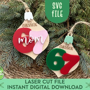 Könnte beinhalten: Zwei Weihnachtsbaumschmuck aus Holz mit dem Text "2025 Survivor" und dem Wort "Mom" in Weiß. Einer hat eine rote "6" und "7" und der andere eine grüne "6" und rote "7". Die Ornamente hängen an einem Bett aus Tannennadeln.