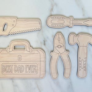 May include: A collection of wooden tool cutouts, including a saw, screwdriver, pliers, hammer, and a toolbox with the text "BEST DAD EVER". The tools are light brown with dark brown outlines, set against a white marble background.