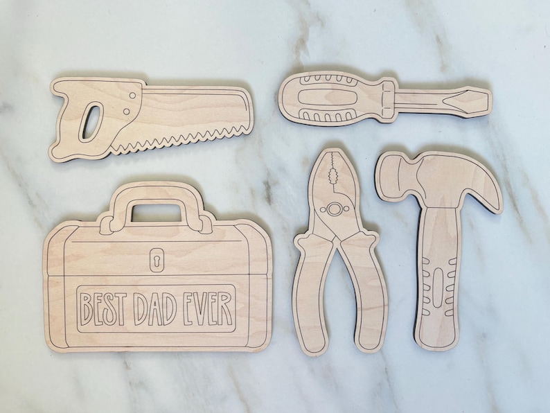 May include: A collection of wooden tool cutouts, including a saw, screwdriver, pliers, hammer, and a toolbox with the text "BEST DAD EVER". The tools are light brown with dark brown outlines, set against a white marble background.