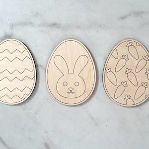 May include: Three wooden Easter egg shapes, each with a different design. One egg has a chevron pattern, another has a bunny face, and the last one has a pattern of carrots.