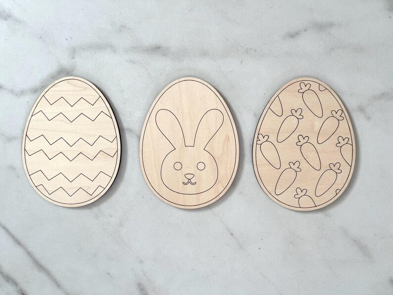 May include: Three wooden Easter egg shapes, each with a different design. One egg has a chevron pattern, another has a bunny face, and the last one has a pattern of carrots.