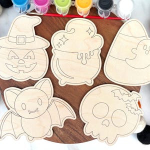 May include: A Halloween-themed craft kit featuring wooden cutouts of a jack-o'-lantern, cauldron, candy corn with a ghost, bat, and skull. The kit includes paint pots and a paintbrush, perfect for a fun DIY project.