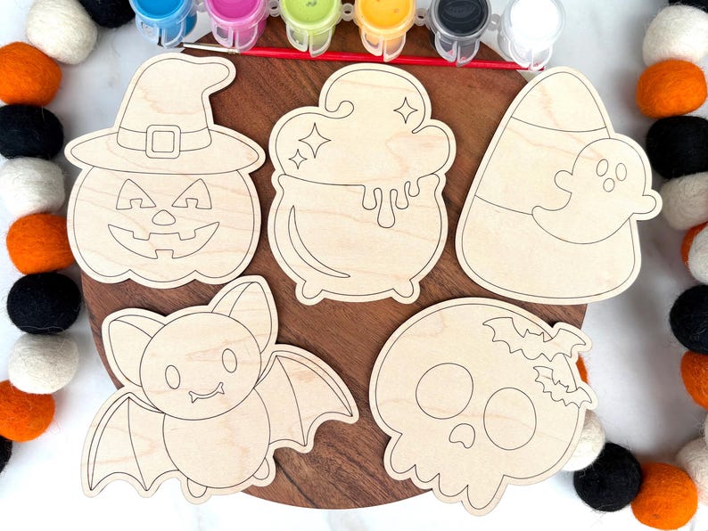 May include: A Halloween-themed craft kit featuring wooden cutouts of a jack-o'-lantern, cauldron, candy corn with a ghost, bat, and skull. The kit includes paint pots and a paintbrush, perfect for a fun DIY project.