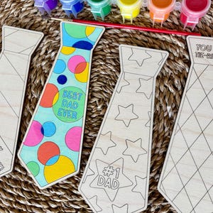 May include: Wooden tie-shaped craft kits with various designs and text, including "Happy Father's Day!", "Best Dad Ever", "#1 Dad", and "You Are Tie-Riffic!". Paint pots and a paintbrush are also visible, suggesting a DIY project.