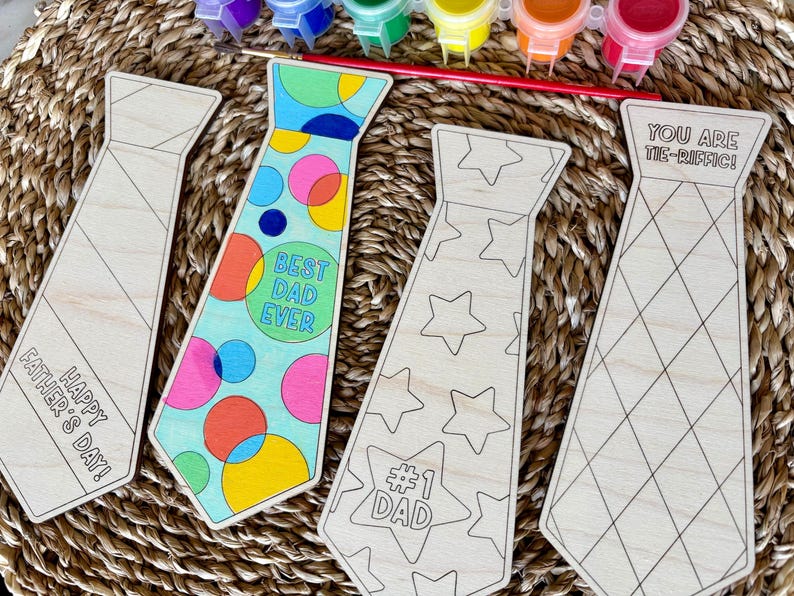 May include: Wooden tie-shaped craft kits with various designs and text, including "Happy Father's Day!", "Best Dad Ever", "#1 Dad", and "You Are Tie-Riffic!". Paint pots and a paintbrush are also visible, suggesting a DIY project.