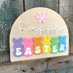 May include: A wooden sign with the words "happy easter" in white script. The sign is decorated with a pink flower and six colourful bunny-shaped figures that spell out "easter".