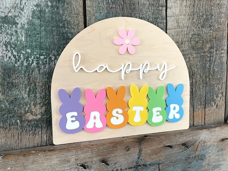 May include: A wooden sign with the words "happy easter" in white script. The sign is decorated with a pink flower and six colourful bunny-shaped figures that spell out "easter".