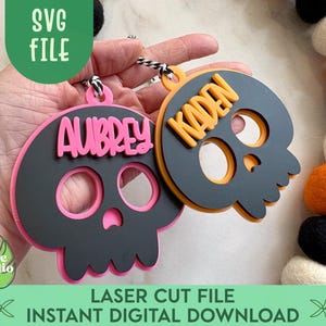 May include: Two skull-shaped ornaments with cut-out eye and nose sockets. One is black with a pink outline and the name "AUBREY" in pink. The other is black with an orange outline and the name "KADEN" in orange. The image also includes the text "SVG FILE" and "LASER CUT FILE INSTANT DIGITAL DOWNLOAD".