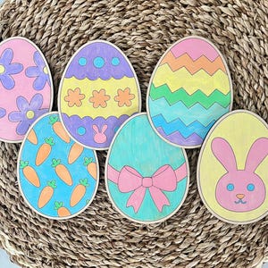 May include: Six wooden Easter eggs painted in various colors and designs. The eggs feature a bunny, carrots, flowers, and geometric patterns.