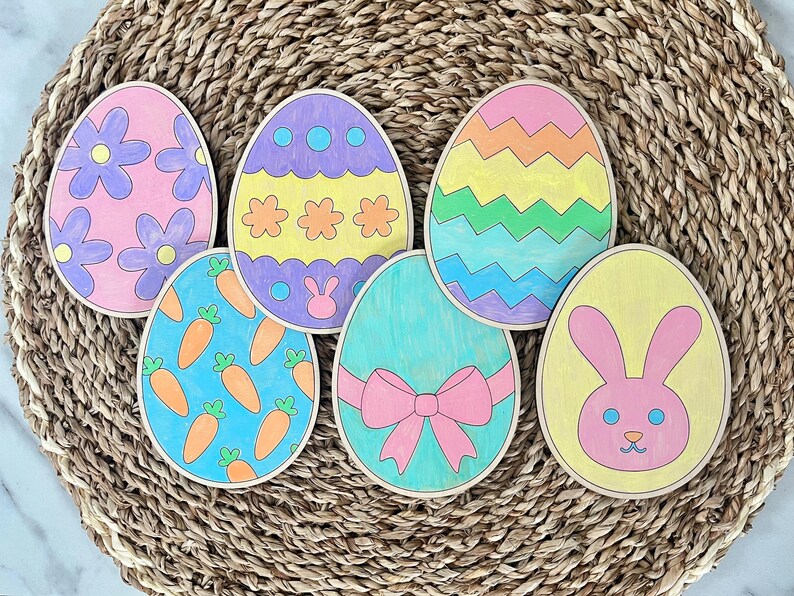 May include: Six wooden Easter eggs painted in various colors and designs. The eggs feature a bunny, carrots, flowers, and geometric patterns.