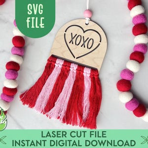 May include: A laser cut wooden heart with the word "XOXO" inside, hanging from a pink and white yarn tassel. The heart is on a white background with a pink and white garland.