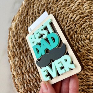 May include: A wooden sign with the words "Best Dad Ever" in teal and green letters. A black paper mustache is layered over the words "Dad".