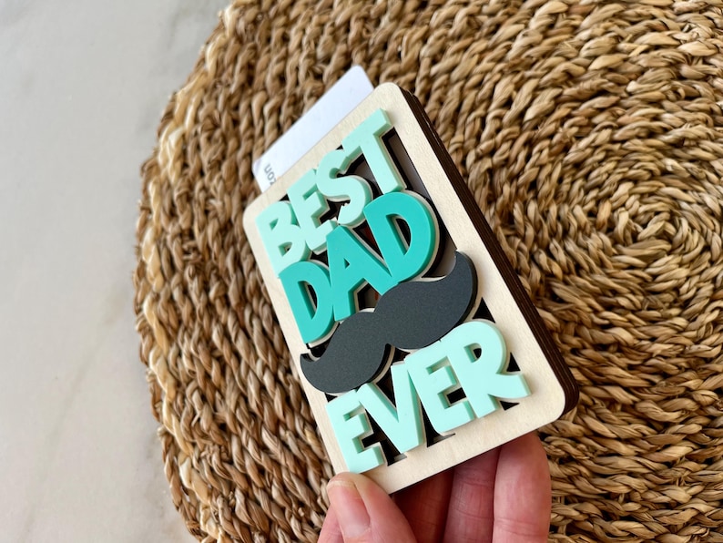 May include: A wooden sign with the words "Best Dad Ever" in teal and green letters. A black paper mustache is layered over the words "Dad".