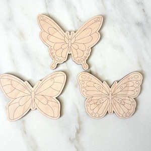May include: Three wooden butterfly cutouts with detailed wing patterns. The butterflies are light brown and appear to be unfinished, ready for painting or staining. The butterflies are arranged on a white marble surface.