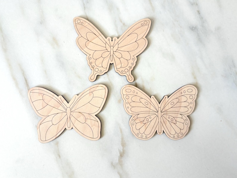 May include: Three wooden butterfly cutouts with detailed wing patterns. The butterflies are light brown and appear to be unfinished, ready for painting or staining. The butterflies are arranged on a white marble surface.