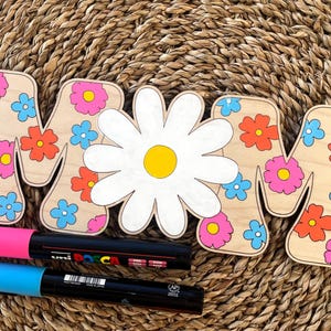 May include: Wooden letters spelling "MOM" decorated with colorful flowers and a large white daisy in the center. The letters are on a woven straw background with two black markers.
