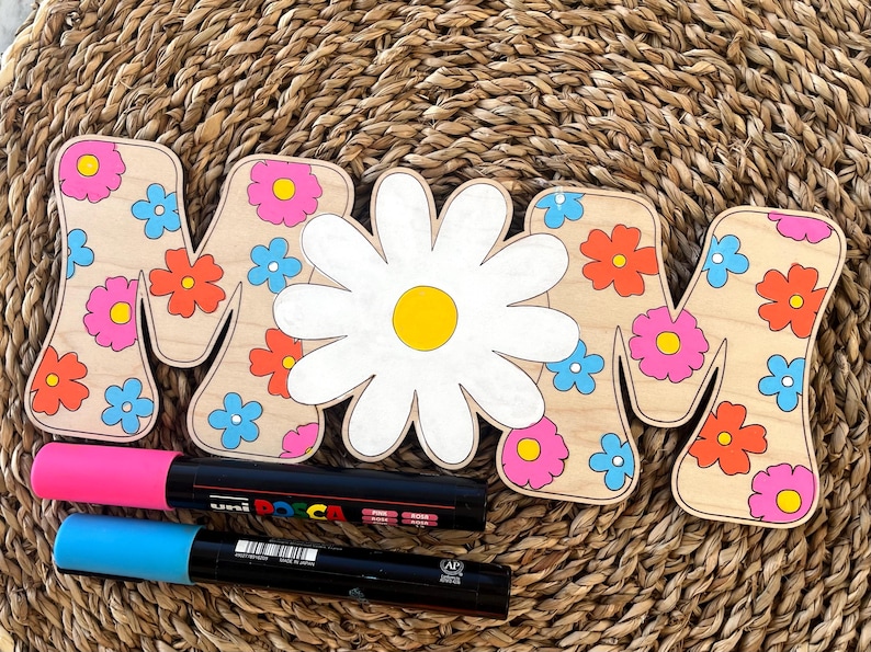 May include: Wooden letters spelling "MOM" decorated with colorful flowers and a large white daisy in the center. The letters are on a woven straw background with two black markers.