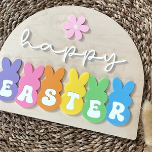 May include: A wooden sign with the words "happy easter" in white script. The sign is decorated with colourful bunny shapes that spell out "easter".