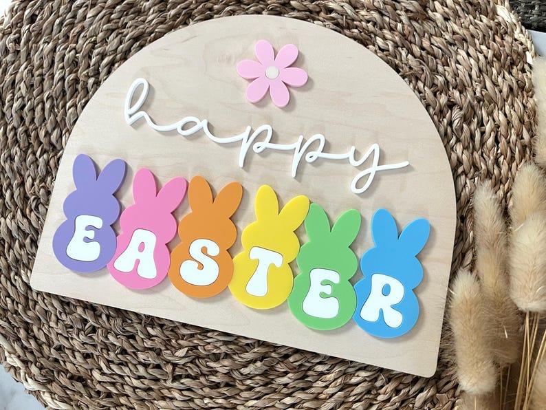May include: A wooden sign with the words "happy easter" in white script. The sign is decorated with colourful bunny shapes that spell out "easter".