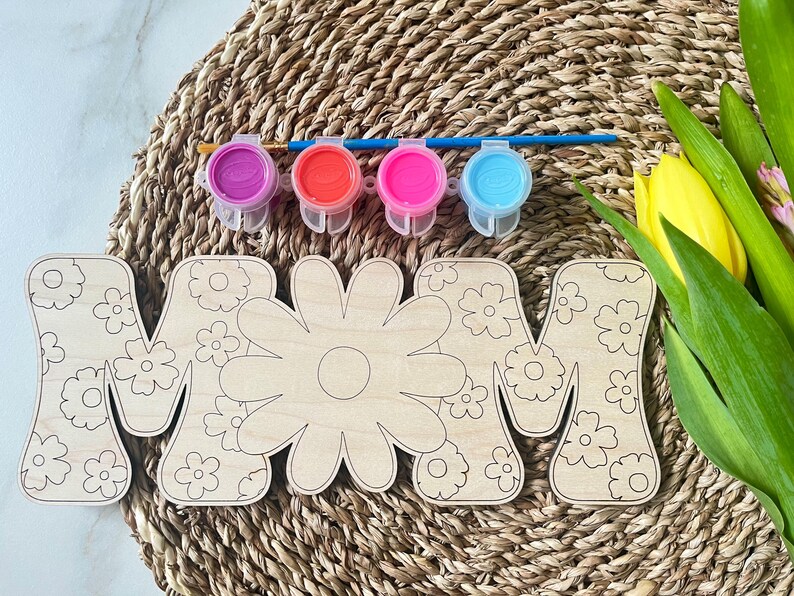 May include: Wooden craft kit featuring the word "MOM" with a floral design. The kit includes four small paint pots in purple, red, pink, and blue.