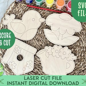 May include: Four unfinished wooden cutouts of a bird, a flower, a birdhouse, and a watering can. The cutouts are on a woven straw mat with paint pots and brushes in the background. The text "SCORE & CUT" and "SVG FILE" are also visible.