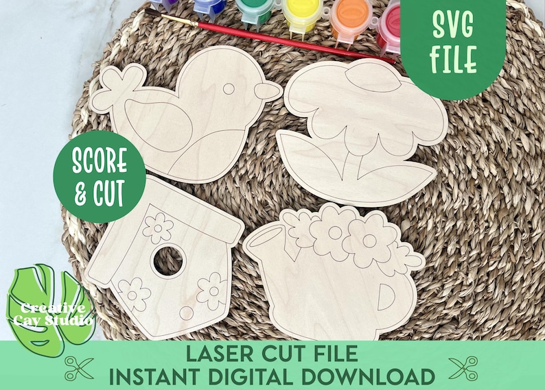 May include: Four unfinished wooden cutouts of a bird, a flower, a birdhouse, and a watering can. The cutouts are on a woven straw mat with paint pots and brushes in the background. The text "SCORE & CUT" and "SVG FILE" are also visible.