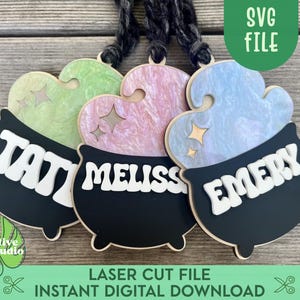 May include: Three black cauldron-shaped ornaments with different colored tops. The tops are pastel green, pink, and blue. Each cauldron has a name written on it in white letters: "Tavi", "Meliss", and "Emery".