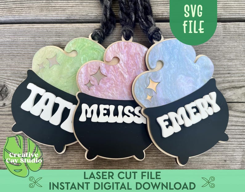 May include: Three black cauldron-shaped ornaments with different colored tops. The tops are pastel green, pink, and blue. Each cauldron has a name written on it in white letters: "Tavi", "Meliss", and "Emery".