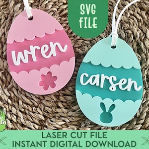 May include: Two pastel Easter egg-shaped ornaments with names "Wren" and "Carsen" written in white. The ornaments are pink and teal with a scalloped edge and a flower and bunny silhouette.