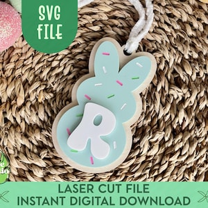 May include: A light blue bunny-shaped Easter cookie with white sprinkles and a white letter "R" in the centre. The cookie is on a woven brown background.