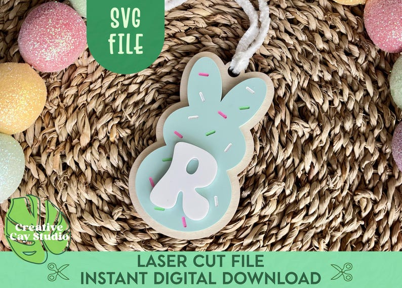 May include: A light blue bunny-shaped Easter cookie with white sprinkles and a white letter "R" in the centre. The cookie is on a woven brown background.