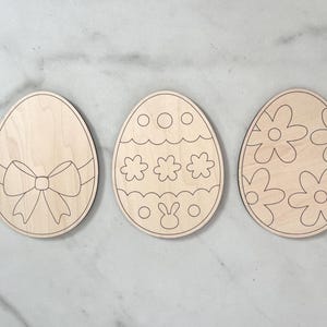 May include: Three unfinished wooden Easter egg shapes. One egg has a bow, one has a bunny and dots, and one has a floral pattern.