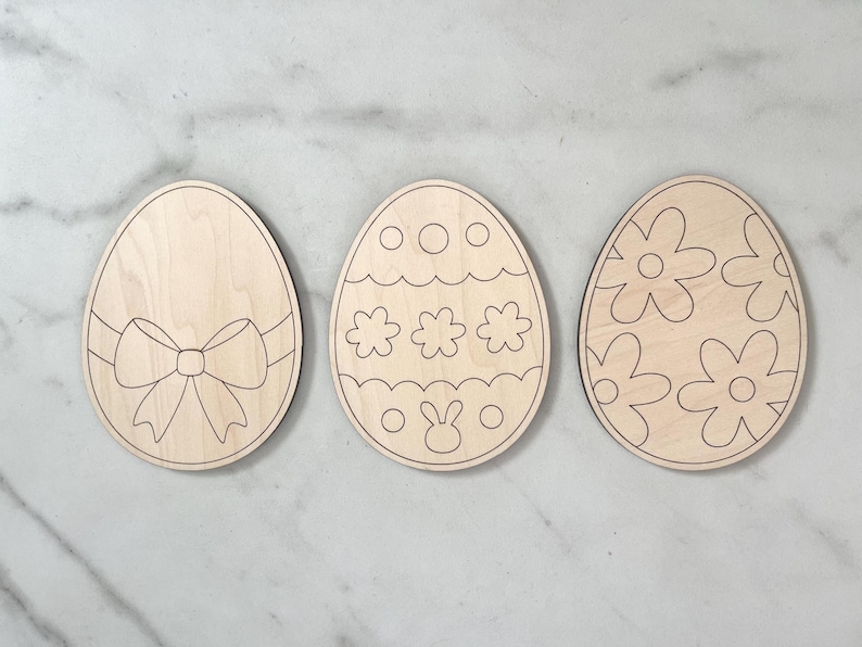 May include: Three unfinished wooden Easter egg shapes. One egg has a bow, one has a bunny and dots, and one has a floral pattern.