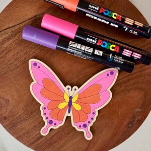 May include: A wooden butterfly with pink, orange, and yellow details sits on a wooden surface, next to three Uni Posca paint markers in orange, pink, and purple. The markers have black barrels and white text.
