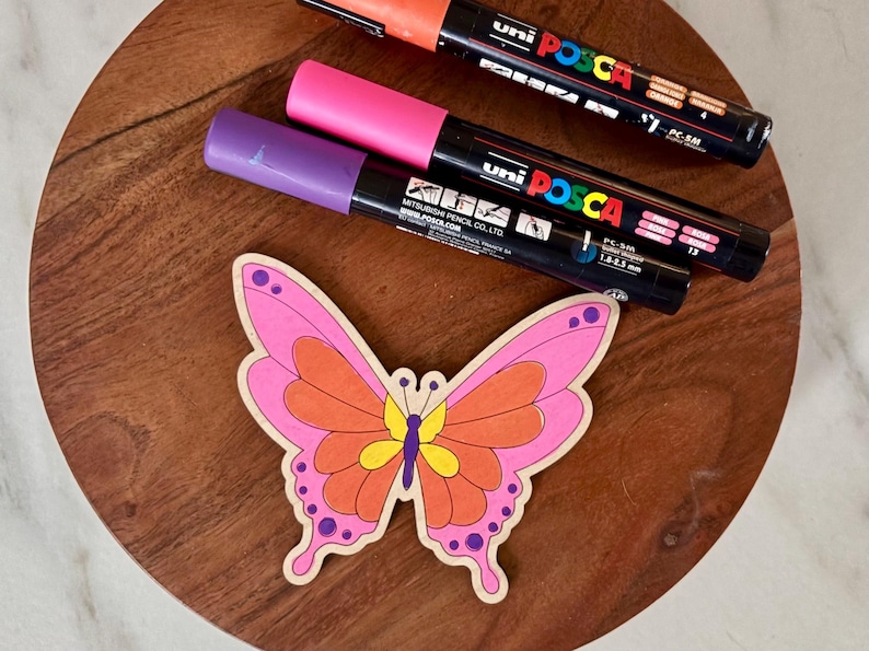 May include: A wooden butterfly with pink, orange, and yellow details sits on a wooden surface, next to three Uni Posca paint markers in orange, pink, and purple. The markers have black barrels and white text.