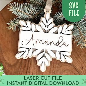 May include: White snowflake ornament with a brown banner that says "Amanda". The ornament is on a wooden surface with pine branches.