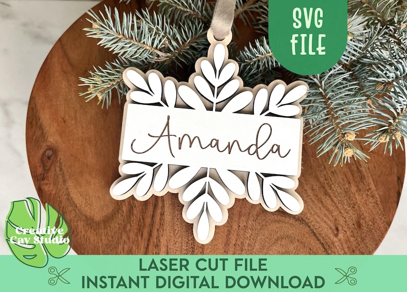 May include: White snowflake ornament with a brown banner that says "Amanda". The ornament is on a wooden surface with pine branches.