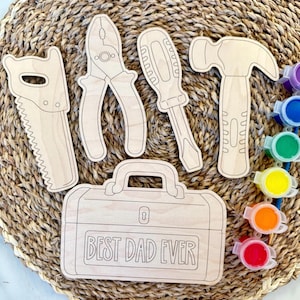 May include: Wooden tool cutouts including a saw, pliers, screwdriver, and hammer, along with a toolbox that reads "BEST DAD EVER." The tools are arranged on a woven surface, with small paint pots in various colors.