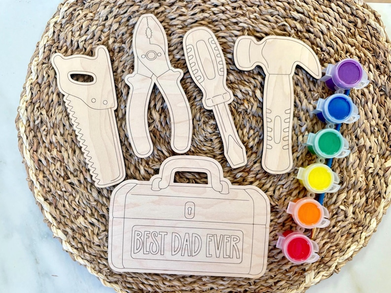 May include: Wooden tool cutouts including a saw, pliers, screwdriver, and hammer, along with a toolbox that reads "BEST DAD EVER." The tools are arranged on a woven surface, with small paint pots in various colors.