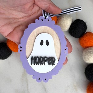 May include: A Halloween-themed wooden ornament featuring a white ghost with black eyes inside a purple frame. The name "HARPER" is displayed below the ghost. The ornament is held by a hand and is part of a garland with black, orange, and white felt balls.