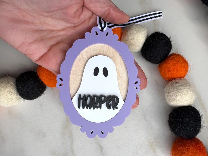 May include: A Halloween-themed wooden ornament featuring a white ghost with black eyes inside a purple frame. The name "HARPER" is displayed below the ghost. The ornament is held by a hand and is part of a garland with black, orange, and white felt balls.