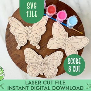 May include: Wooden butterfly cutouts arranged on a wooden surface, with paint pots and a paintbrush. The image includes text that reads "SVG FILE", "SCORE & CUT", "LASER CUT FILE", and "INSTANT DIGITAL DOWNLOAD".