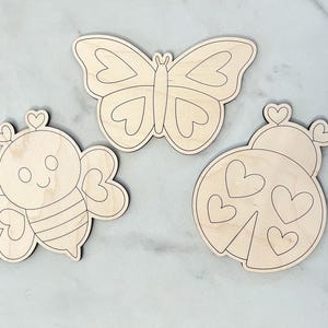 Puede incluir: Tres formas de madera sin terminar recortadas de una mariposa, una abeja y una mariquita. Cada forma tiene un dise&ntilde;o de coraz&oacute;n.