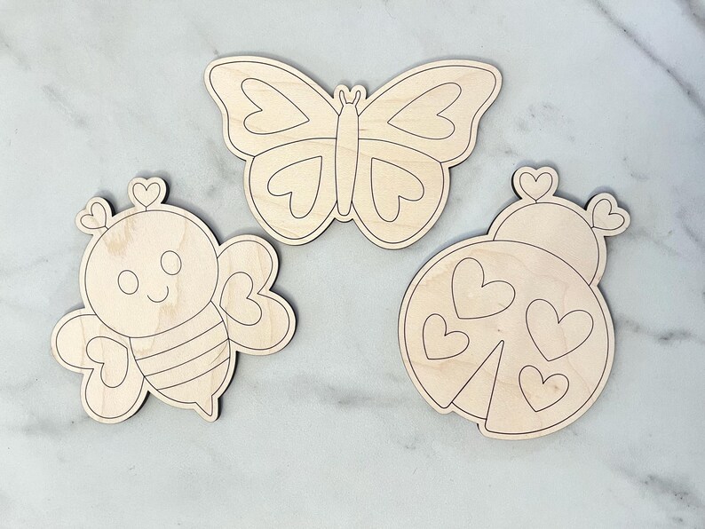 Puede incluir: Tres formas de madera sin terminar recortadas de una mariposa, una abeja y una mariquita. Cada forma tiene un dise&ntilde;o de coraz&oacute;n.