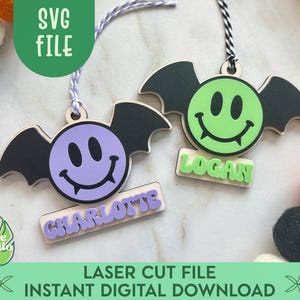 May include: Two wooden bat-shaped ornaments with smiley faces and personalized name tags. One ornament has a purple face and the other has a green face. The text "SVG FILE" and "LASER CUT FILE INSTANT DIGITAL DOWNLOAD" are also visible.